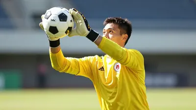 A professional action close-up shot of a goalkeeper in a yellow jersey catching a football on a sunlit grass, blurred...