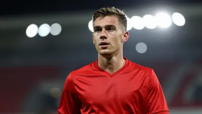 A young male substitute in his early twenties, wearing a solid red Sarpsborg 08 jersey, captured waist-up with a focused...