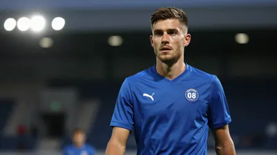 A mid-30s anonymous Sarpsborg 08 midfielder in a solid blue kit, waist-up, focused expression, background blurred stadium...